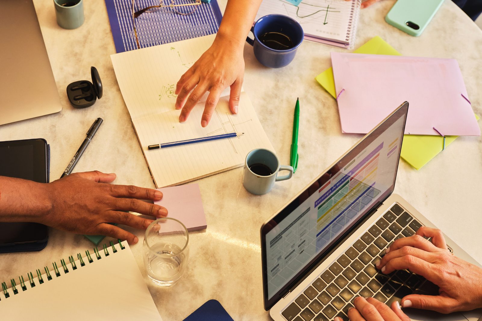 Hands at a shared desk covered with notebooks, a laptop and coffee cups during a working session